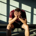 AI-generated image: Dramatic overhead angle of a young athlete wearing a burgundy training shirt sitting alone on a gym